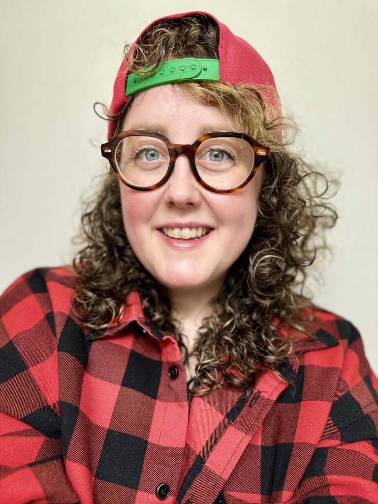 Amy smiles at the camera, she wears a red and black flannel shirt and a Super Sonics snapback hat